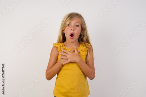 Scared Little girl with beautiful blonde hair over white wall looks with frightened expression, keeps hands on chest, being puzzled to notice something strange,  People, hush reaction and emotions.