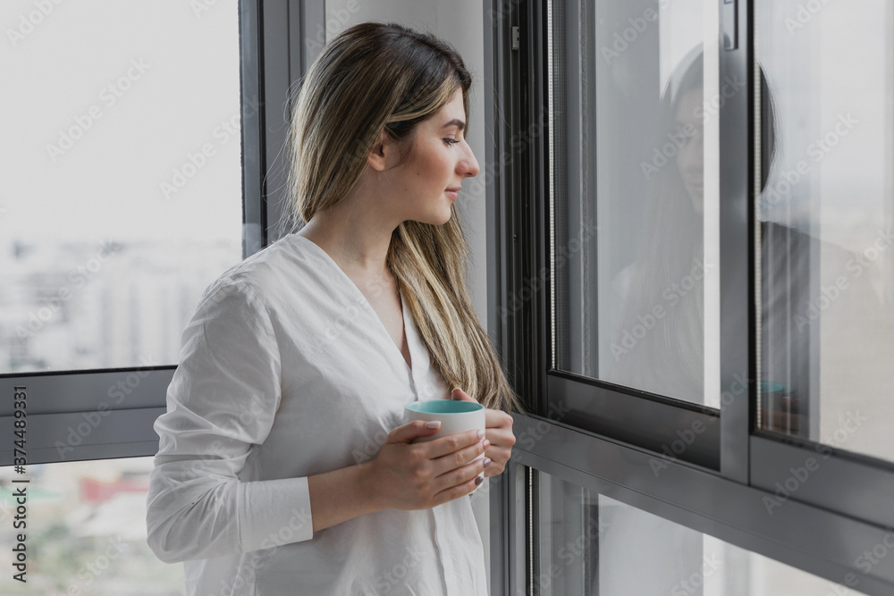 Young beautiful woman near window with cup of coffee or tea at home. Dream and relax. Good day. Woman in the morning is holding a mug and looking at her child in the playground from home. Young beautiful woman near window with cup of coffee or tea at home. Dream and relax. Good day. Woman in the morning is holding a mug and looking at her child in the playground from home.