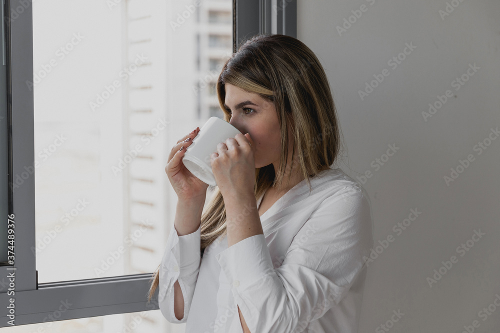 Young beautiful woman near window drinking cup of coffee or tea at home. Dream and relax. Good day. Woman in the morning is holding a mug and looking at the sunrise and having a perfect cozy morning Young beautiful woman near window drinking cup of coffee or tea at home. Dream and relax. Good day. Woman in the morning is holding a mug and looking at the sunrise and having a perfect cozy morning