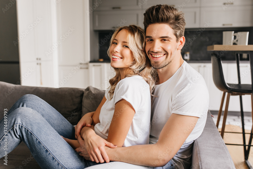 Smiling attractive couple sitting on sofa hugging each other and looking to camera. Two young happy people share morning together. Smiling attractive couple sitting on sofa hugging each other and looking to camera. Two young happy people share morning together.
