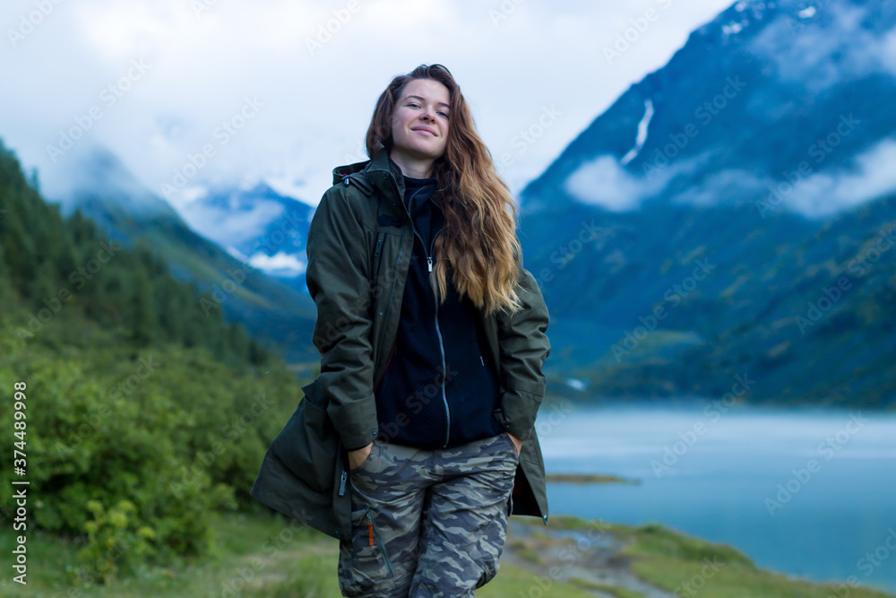 Young woman in a jacket and trousers against the backdrop of mountains. Girl tourist in warm clothes at dawn against the background of fog, lake and blue mountains.