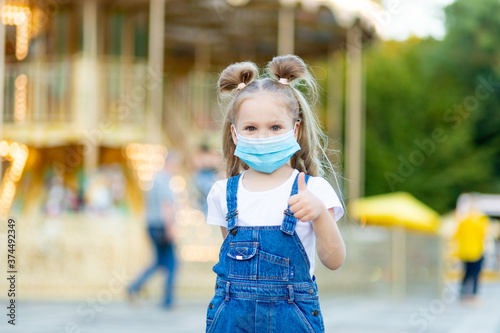 a baby girl stands in an amusement Park wearing a protective medical mask and gives a thumbs up