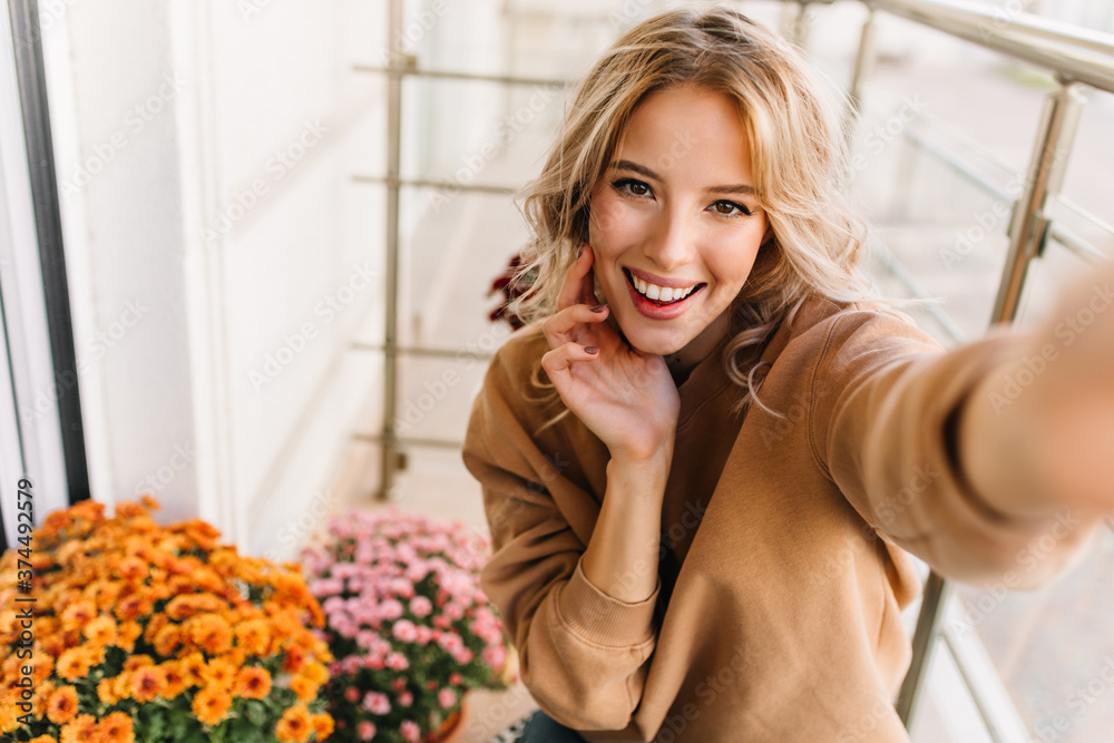 Romantic fair-haired girl posing at balcony with pretty smile. Enchanting young lady making selfie with orange flowers. Romantic fair-haired girl posing at balcony with pretty smile. Enchanting young lady making selfie with orange flowers.
