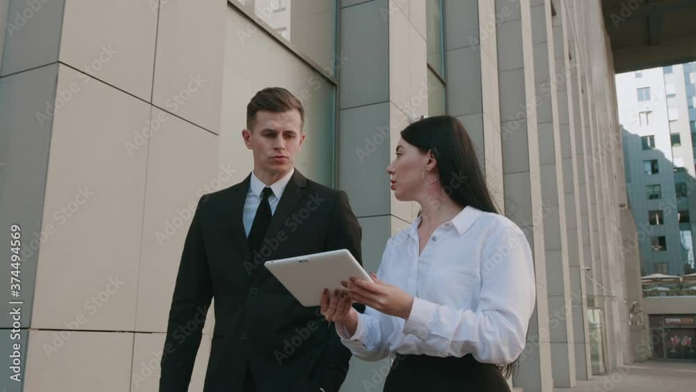 Businessman and businesswoman are walking downtown and discussing business on background of office building. Colleagues man and woman on the way to work are discussing startup with using tablet Businessman and businesswoman are walking downtown and discussing business on background of office building. Colleagues man and woman on the way to work are discussing startup with using tablet
