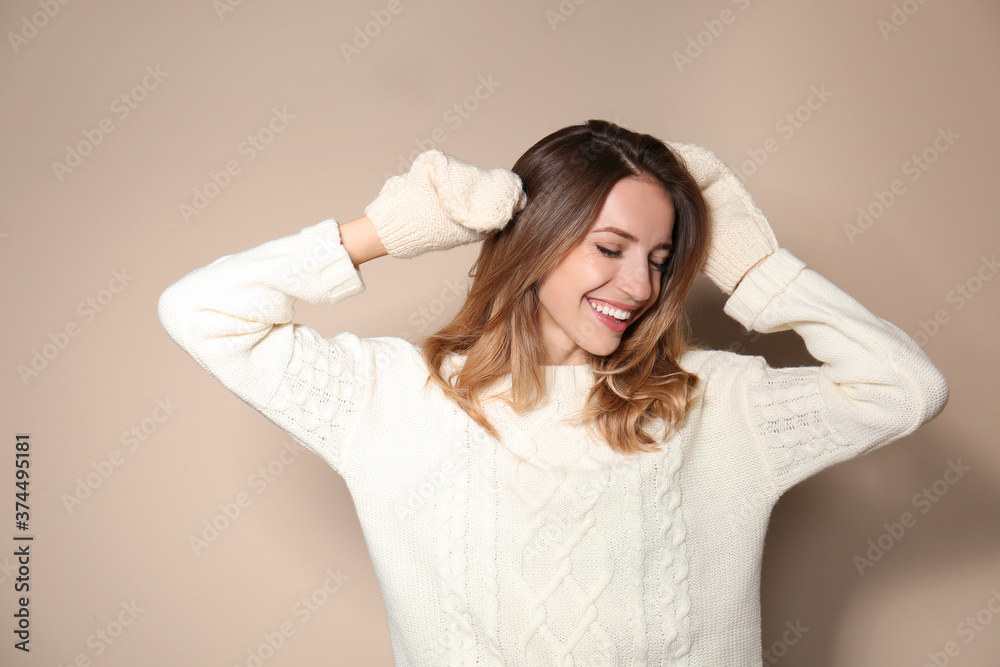 Happy young woman wearing warm sweater and knitted mittens on beige background