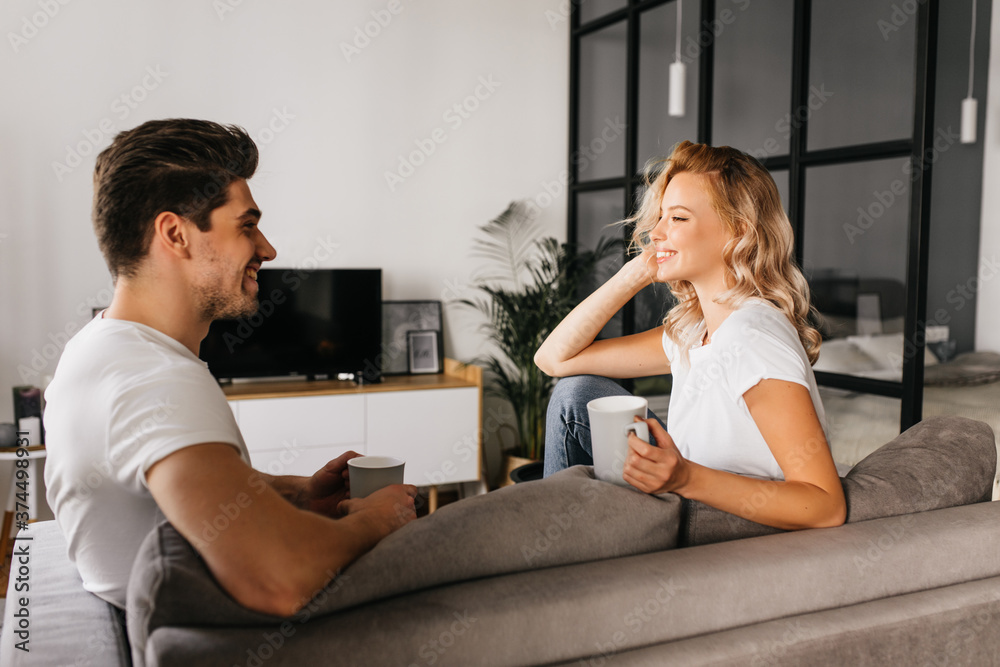 Smiling young couple sitting at home looking at each other and holding cups. Cozy home photo of two attractive people in love spending time together. Smiling young couple sitting at home looking at each other and holding cups. Cozy home photo of two attractive people in love spending time together.