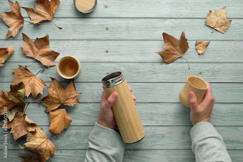 Zero waste coffee in bamboo cups from eco friendly insulated metal flask in Fall. Hands holding the flask and cup. Top view on aged blue mint wooden background with dry Autumn sycamore leaves. photo