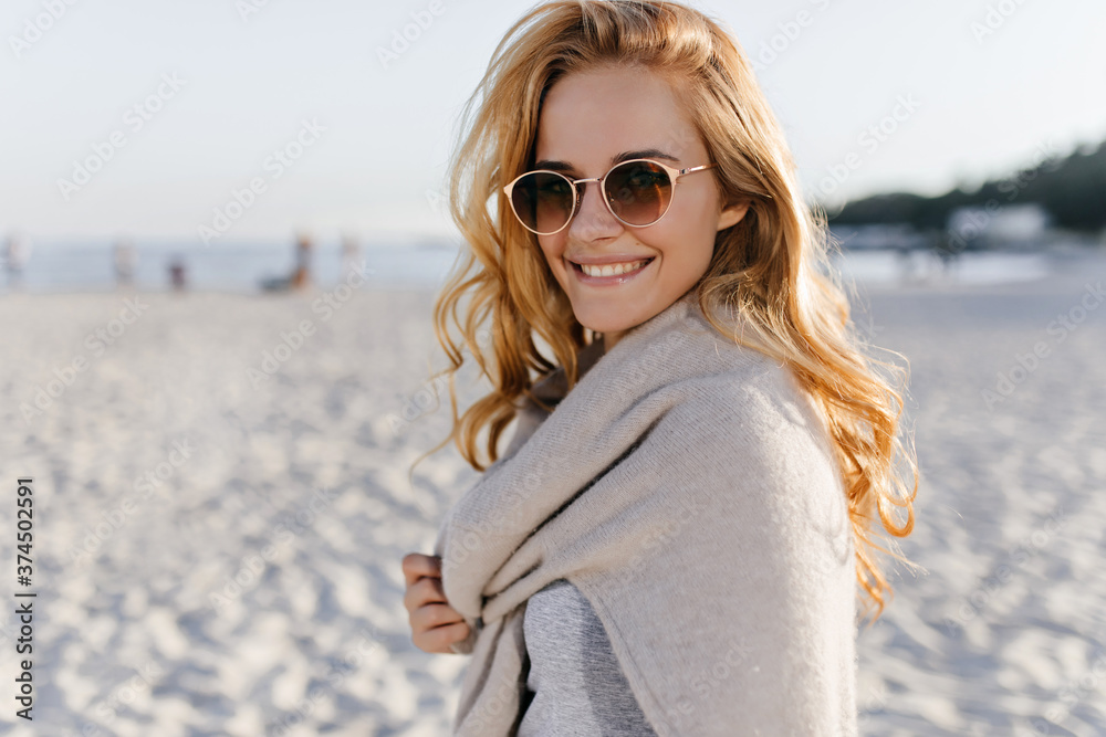 Snapshot of curly young woman in beige outfit and sunglasses smiling against backdrop of sea Snapshot of curly young woman in beige outfit and sunglasses smiling against backdrop of sea