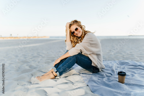 Well-dressed young woman posing with pleasure at sea in cold autumn day. Outdoor portrait of pleasant blonde girl sitting on sand