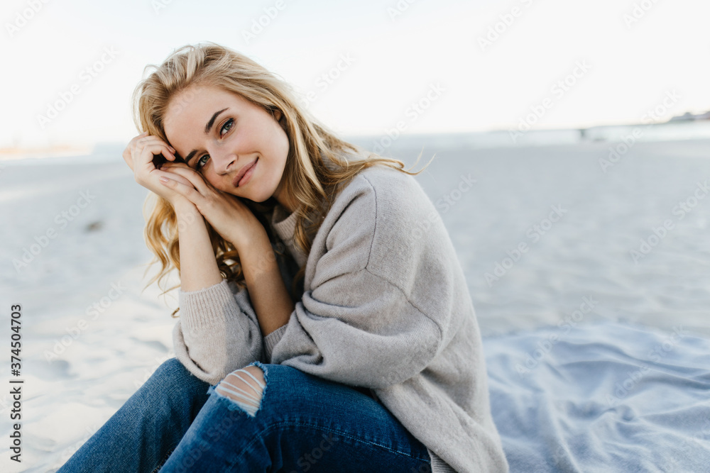 Green-eyed woman in jeans and sweater is sitting on sand, leaning on her knees and looking into camera against background of sea Green-eyed woman in jeans and sweater is sitting on sand, leaning on her knees and looking into camera against background of sea
