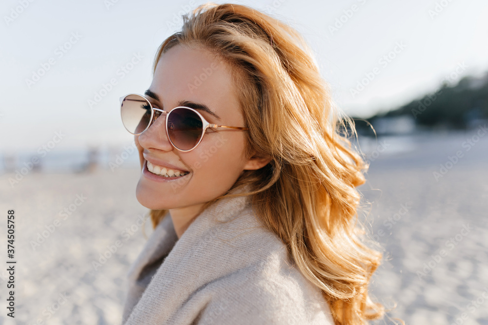 Close-up portrait of positive girl with wavy blonde hair dressed in beige cashmere sweater and sunglasses on beach Close-up portrait of positive girl with wavy blonde hair dressed in beige cashmere sweater and sunglasses on beach