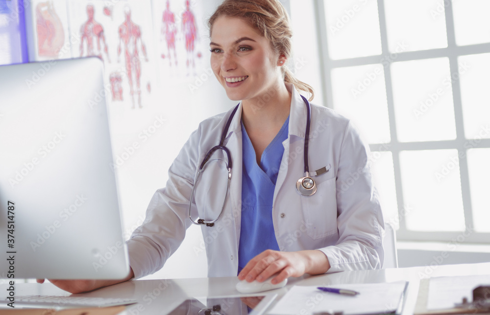 Young female doctor sitting at a desk and working on the computer at the hospital office. Health care, insurance and help concept. Physician ready to examine patient Young female doctor sitting at a desk and working on the computer at the hospital office. Health care, insurance and help concept. Physician ready to examine patient