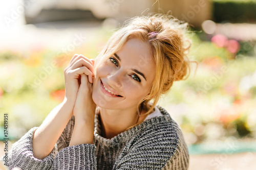 Charming curly girl in knitted sweater looks into camera with smile, posing in garden