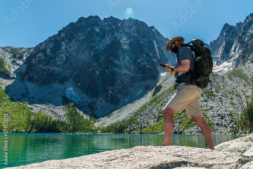 Hiker wearing face mask checking smart phone device at the shore of a gorgeous alpine lake under spectacular mountain. 