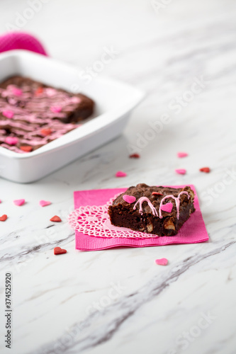 Fototapeta Naklejka Na Ścianę i Meble -  Pan of Homemade Brownies with Pink Frosting on a White Countertop with Heart-Shaped Sprinkles; Piece on Pink Napkin and White Heart-Shaped Doily in Front with Heart-Shaped Sprinkles Scattered Around