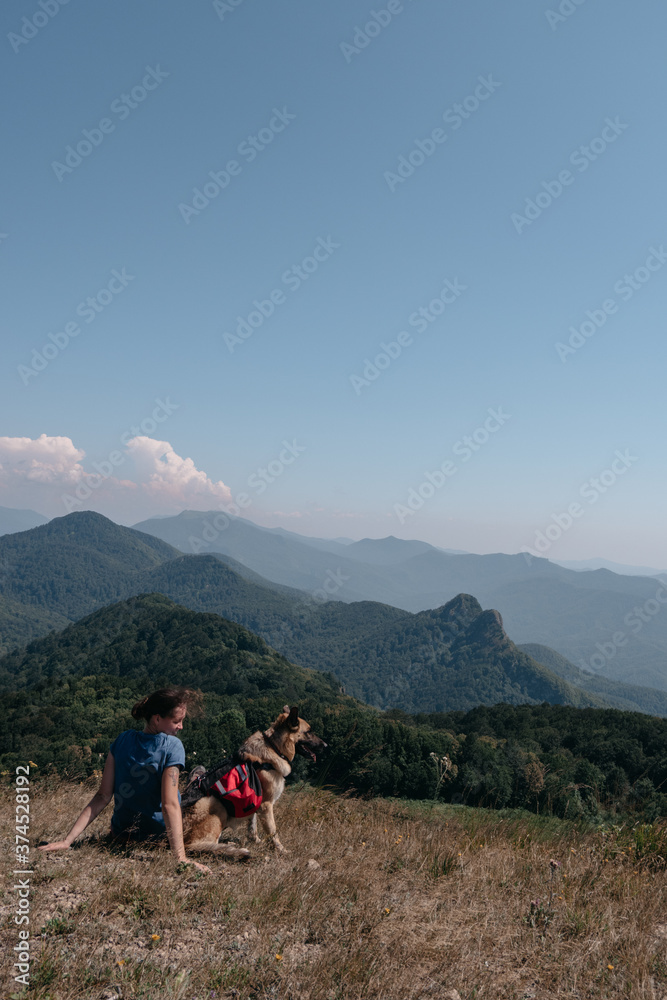 Fototapeta premium Dog traveler goes to the mountains with his best friend man and enjoys nature and beautiful scenery. A young traveller girl is sitting on top of a mountain with her German shepherd.