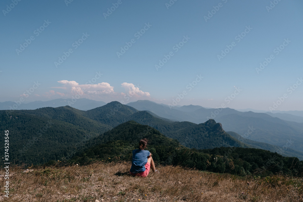 Fototapeta premium Beautiful mountain and hilly landscape in warm, Sunny weather and the young tourist. A female traveler sits on top of a hill and looks at the surrounding mountains.