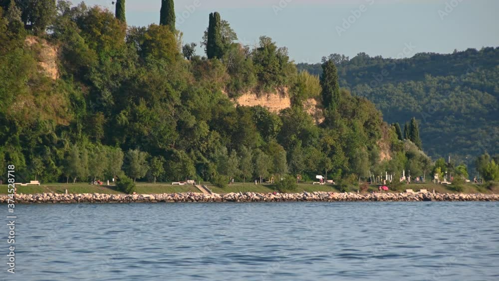 Beautiful coast of Slovenia on bright sunny day. People enjoying at shore on summer day. Trees growing on cliff. Wide, static shot Beautiful coast of Slovenia on bright sunny day. People enjoying at shore on summer day. Trees growing on cliff. Wide, static shot