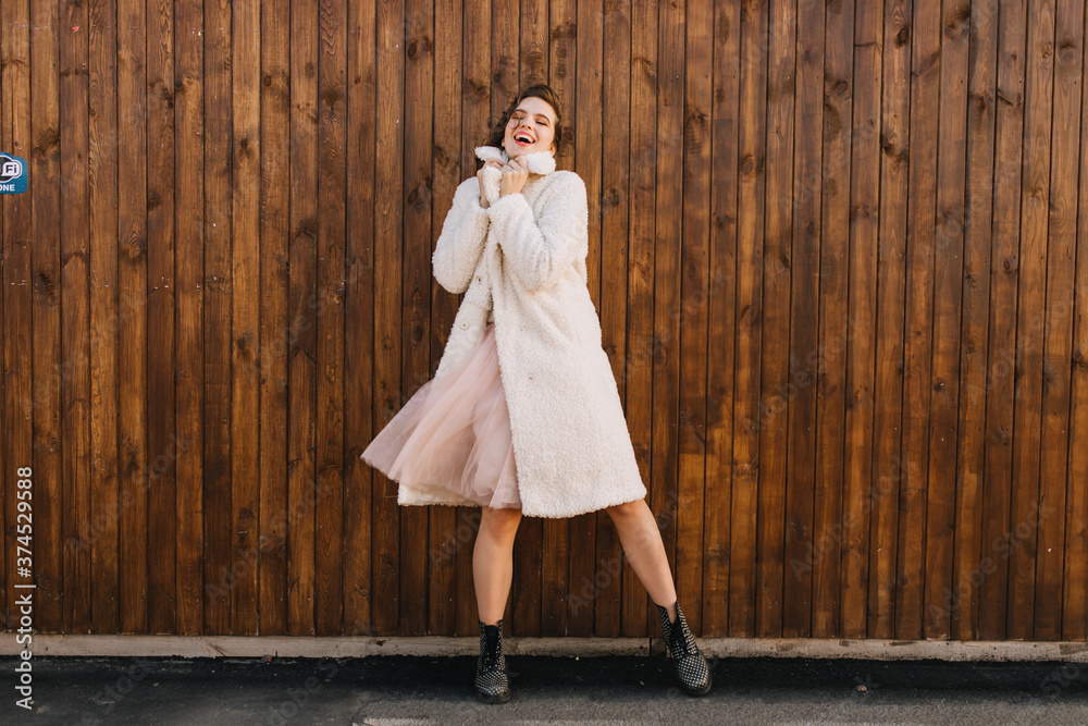Full-length shot of laughing carefree girl dancing on wooden background. Outdoor portrait of pretty european woman in pink long coat.