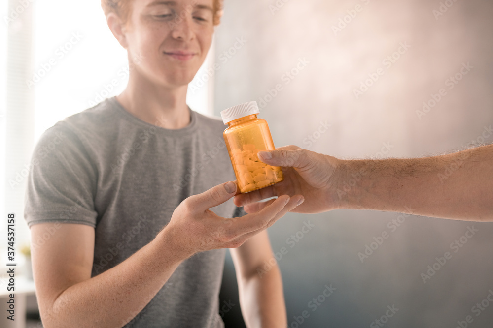 Hand of contemporary physiotherapist passing bottle of pills to young patient Hand of contemporary physiotherapist passing bottle of pills to young patient