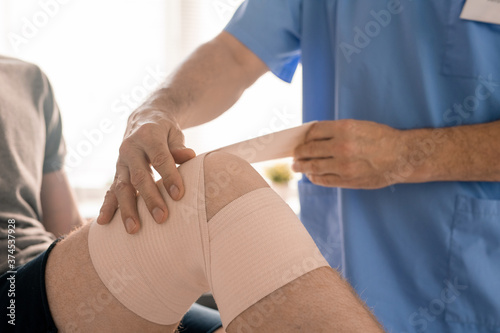 Hands of clinician in blue uniform wrapping injured knee of patient with bandage