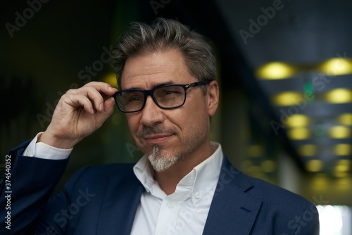 Portrait of happy mature man wearing glasses. Confident businessman wearing suit, standing in office building hallway. Dark, warm colors.
