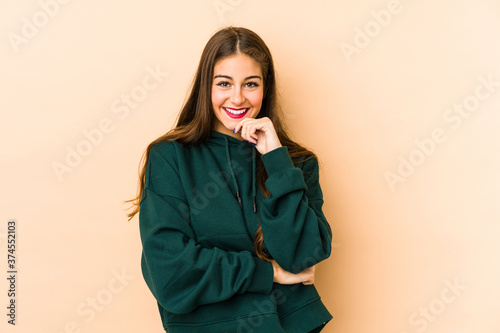 Young caucasian woman isolated en beige background smiling happy and confident, touching chin with hand. © Asier