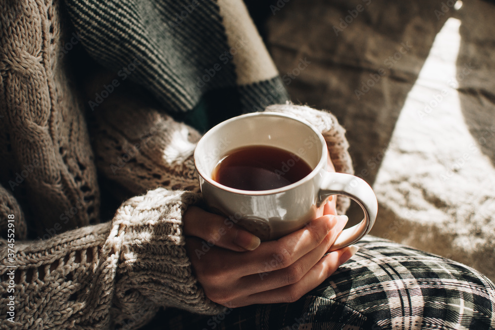 Cozy autumn or winter atmosphere. Young woman with cup of tea and warm sweater is seating at home near the window. Scandinavian hygge concept. Cozy autumn or winter atmosphere. Young woman with cup of tea and warm sweater is seating at home near the window. Scandinavian hygge concept.