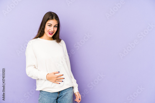 Young caucasian woman isolated on purple background touches tummy, smiles gently, eating and satisfaction concept.