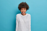 Beautiful happy African American woman has surprised joyful expression, cannot believe sudden success, dressed in casual white jumper, isolated on blue background. Positive reaction concept.