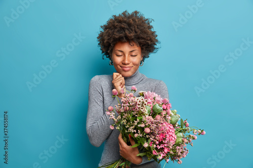 Satisfied Afro American woman receives flower delivery on birthday, satisfied by pleasant present, enjoys holiday greetings, dressed in grey turtleneck, isolated over blue wall. Celebrating womens day