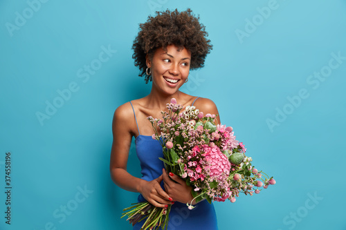 Happy teacher poses with big beautiful bouquet of flowers received from pupils, looks gladfully aside, adores fresh pleasant aroma, enjoys favourite fragrance, wears blue dress, stands indoor.