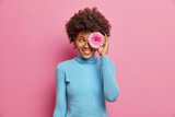 Happy positive dark skinned young woman covers eye with pink gerbera, has toothy smile, dressed casually, poses indoor, enjoys spring time. Flower arranngement. Fashion style. Pink background.
