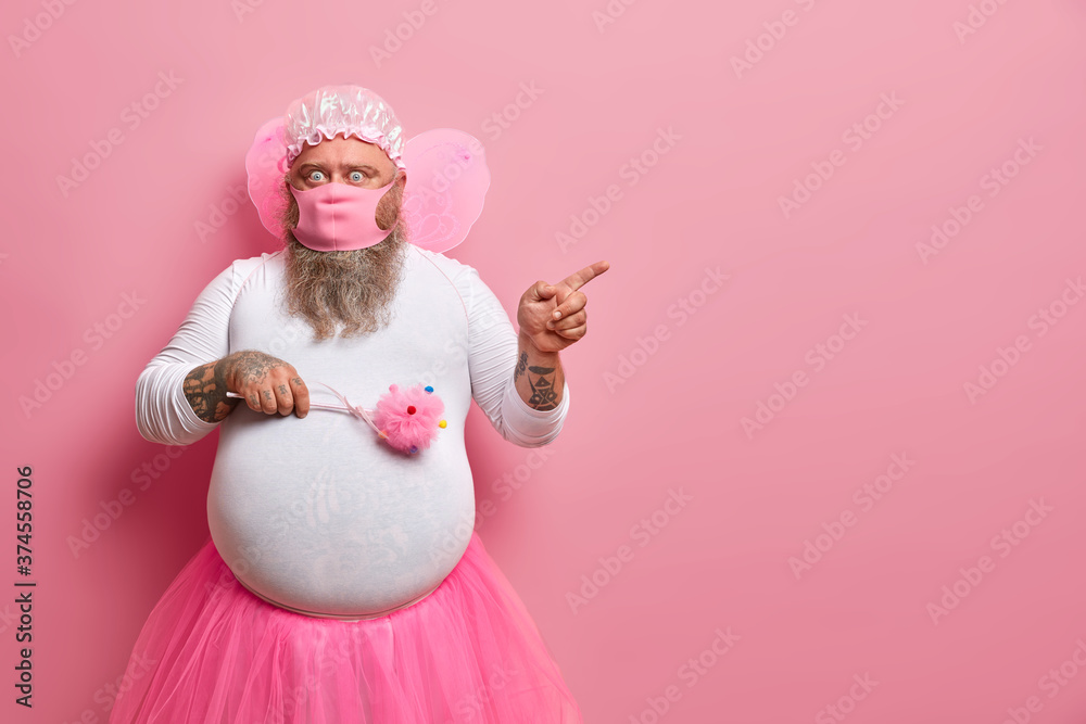 Fototapeta premium Surprised man foolishes around while staying at home during quarantine for long period of time, wears protective mask, fairy costume, indicates on copy space, shows stunning thing, rosy background
