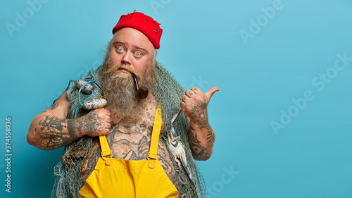 Bearded fisher catches fish with net, enjoys fishing hobby and his occupation, poses with pipe in mouth, points thumb away, shows blank empty space for your advertisement or promotional text