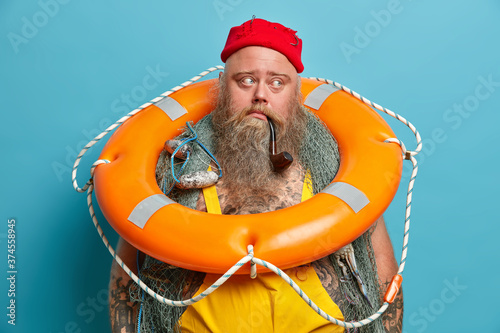 Thoughtful mariner carries inflated ring buoy on neck, ready to rescue people at sea, has thick long beard, smokes pipe, contemplates about something. Seafarer with safety equipment stands indoor