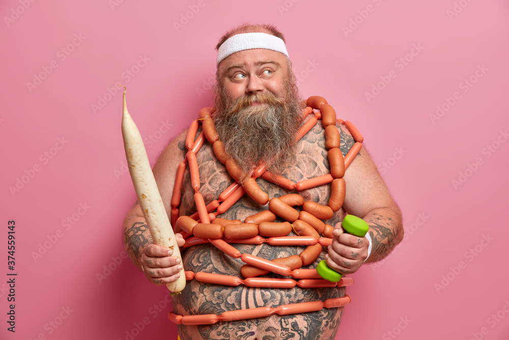 Obesity and proper nutrition concept. Overweight bearded man with naked tattooed body, holds big white radish, wrapped with sausages, busy doing fitness exercises, isolated on pink background.