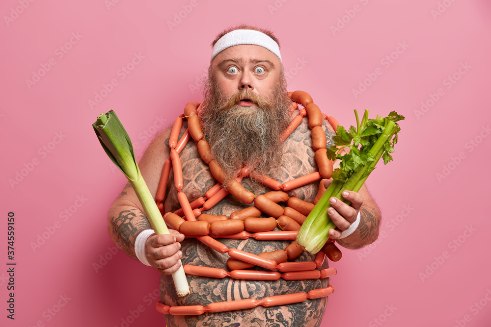 Photo of obese man with thick beard eats useful vegetables, has excess weight, tries to refuse eating sausages, has eyes popped out, has healthy nutrition to keep fit, wears headband, poses indoor