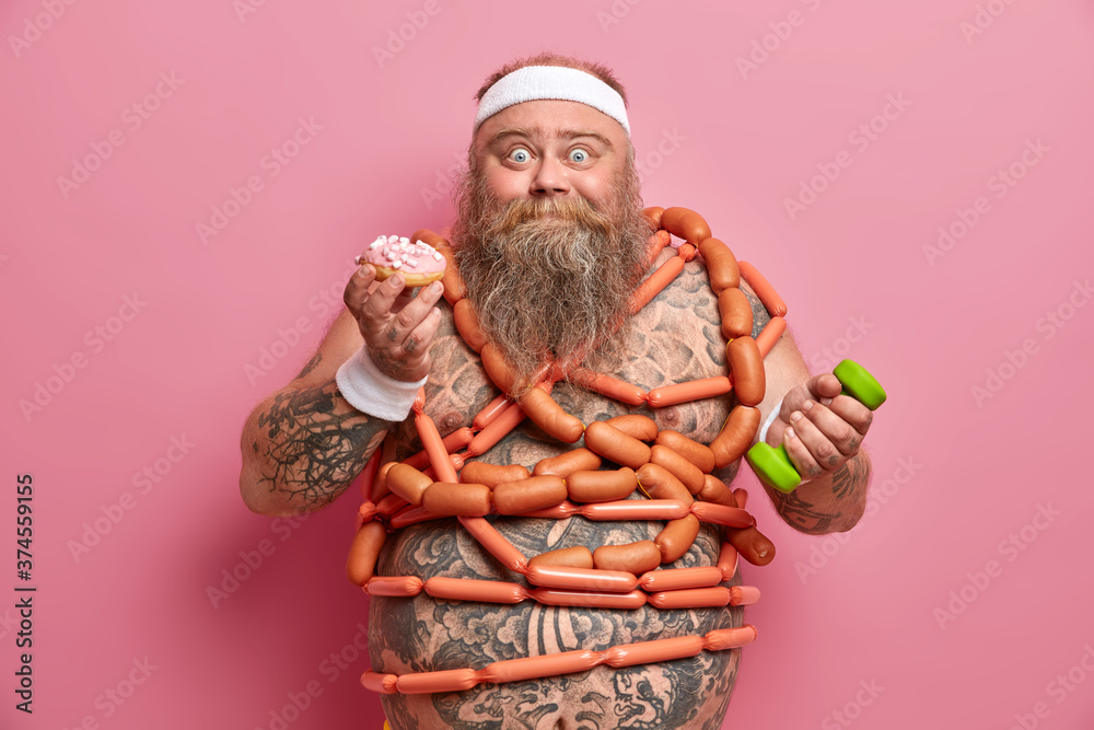 Obesity causes. Funny bearded adult man with excess weight, struggles against sugar addiction, eats donut and sausages, has unproper nutrition, raises dumbbell, has tattooed belly, poses indoor