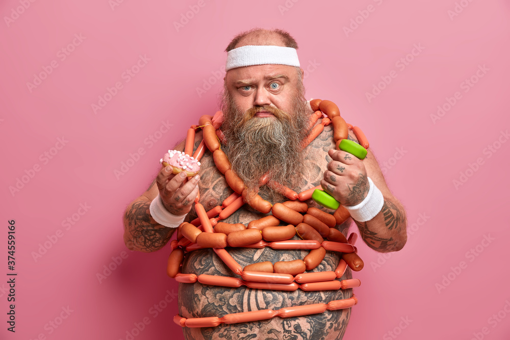 Weight loss, dieting and fitness concept. Serious bearded fatty man holds high calorie sweet donut, refuses eating junk food, goes in for sport regularly, has big belly wrapped with sausages
