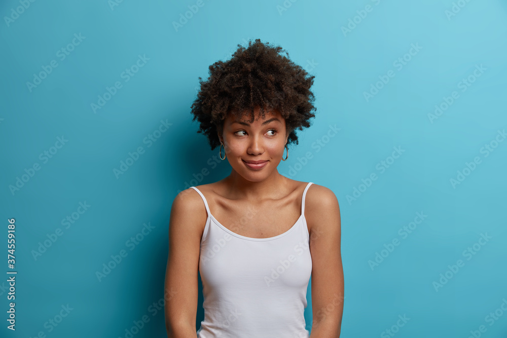 Dreamy positive dark skinned woman with Afro hair looks aside, has pleasant smile on face, dressed in casual vest, notices something pleasant, poses against blue background. Human face expressions Dreamy positive dark skinned woman with Afro hair looks aside, has pleasant smile on face, dressed in casual vest, notices something pleasant, poses against blue background. Human face expressions