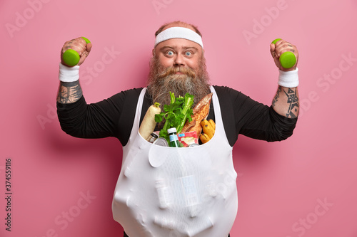 Foto Serious overweight man poses with food products, raises dumbbells, does sport exercises, struggles against excess weight, wears headband and wristband, isolated on pink background
