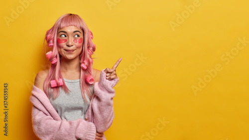Horizontal shot of pink haired woman points aside on yellow background, advertises product, wears collagen patches under eyes, hair curlers, makes hairstyle, promots item for caring about yourself