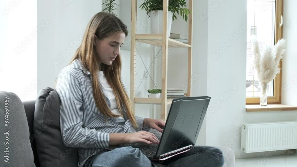 smiling young woman sitting on couch, using laptop notebook, looking at screen, typing message, happy lady chatting on computer, browsing, surfing internet, studying or working online at home