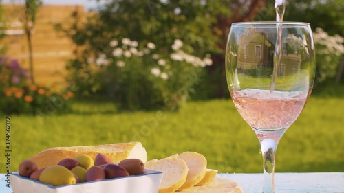 Close up rose wine pouring into a glass from a bottle. Bread and olives on the table in the garden of country house. Summer day, blurry natural background. Macro shot with slide or move camera