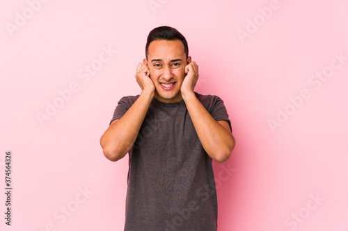 Yooung latin man posing in a pink backgroundcovering ears with hands.