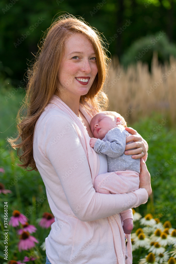 Medium vertical shot of pretty smiling red-haired young woman holding her newborn baby girl tenderly while standing in blooming garden Medium vertical shot of pretty smiling red-haired young woman holding her newborn baby girl tenderly while standing in blooming garden