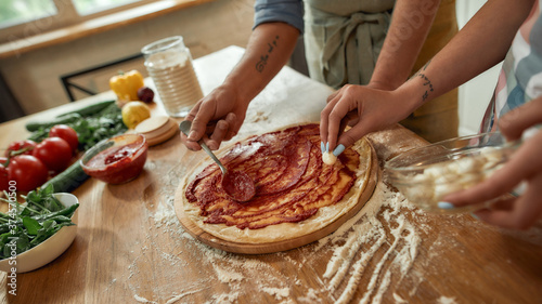 Team work. Cropped shot of couple making pizza together at home. Man in apron adding, applying tomato sauce on the dough while woman adding mozzarella cheese. Hobby, lifestyle