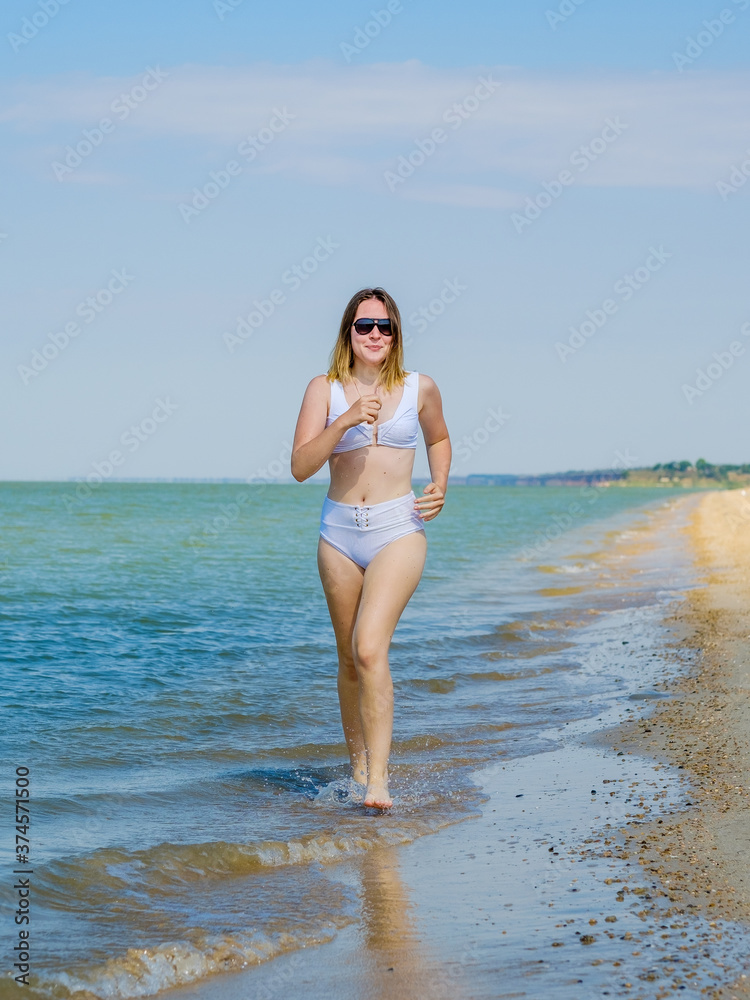 A young girl runs along the sandy sea beach along the surf. Splashes of sea water. The girl is wearing a white swimsuit and sunglasses. Freedom and carelessness. Outdoor fitness. Sunny day. Copy space A young girl runs along the sandy sea beach along the surf. Splashes of sea water. The girl is wearing a white swimsuit and sunglasses. Freedom and carelessness. Outdoor fitness. Sunny day. Copy space
