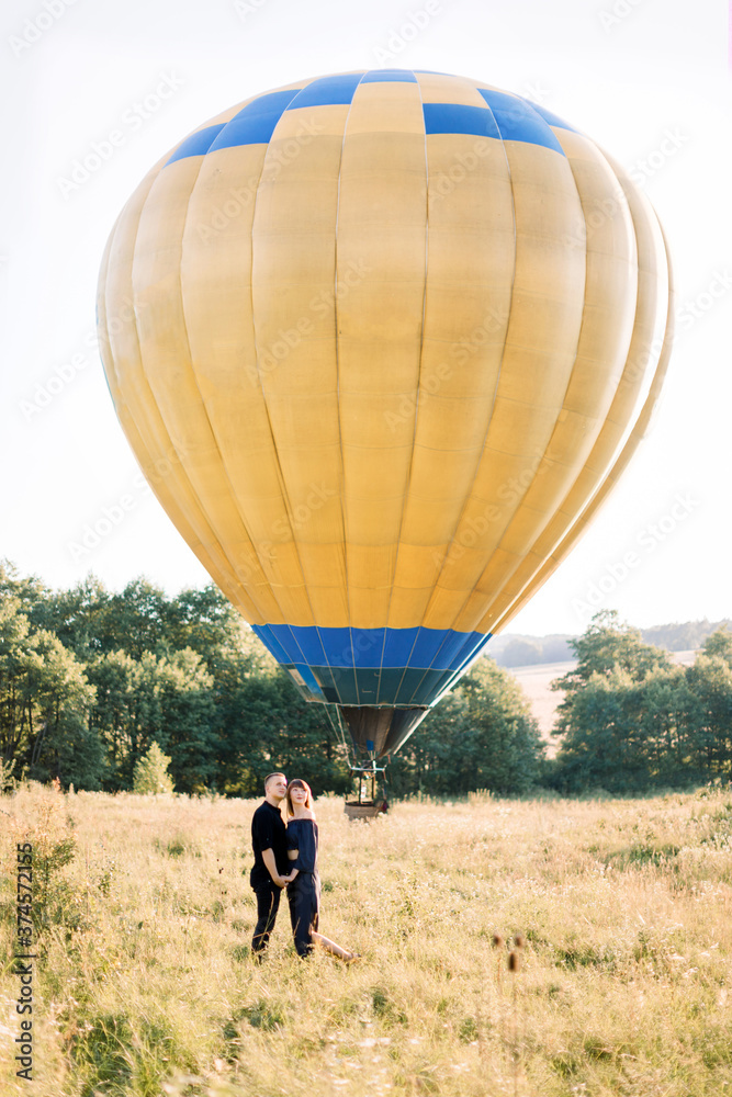 Obraz premium Full length portrait of lovely young couple in black, hugging each other and enjoying summer walk in the field, waiting for their air balloon tour. Yellow air balloon on the background